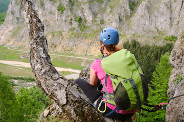 Woman high up on a via ferrata route called 'Spirala Mare' in Baia de Fier, Gorj county, Romania, gazes in the distance, sitting down next to a tree trunk.
