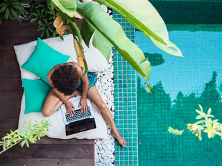 bird view of a remote online working digital nomad women with curly hair and laptop sitting crossed legged at a sunny turquoise water pool surrounded by cushions and plants in the foreground