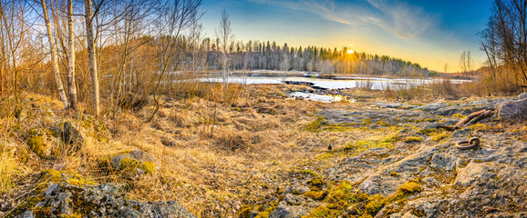 An outdoor scenic beauty of sunset through the woods with golden light on the grass and half frozen snow lake