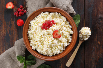 Fermented milk product. Homemade cottage cheese in a clay bowl with berries: strawberries, red currants, mint and a spoon on an old wooden background. Flatlay, top view. Background image, copy space