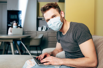 selective focus of freelancer in medical mask near laptop and girlfriend at home