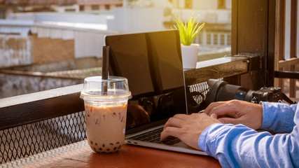 A young businessman also works in a coffee shop, with a notebook computer placed on a table with a camera and a glass of milk tea with a blurred background.