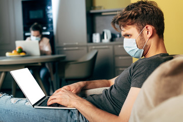 selective focus of freelancer in medical mask using laptop with white screen near girlfriend at home