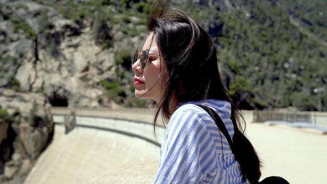 beautiful asian young girl backpacker relying leaning on railing of bridge enjoy wind blowing hair on sunny cool day. curiously woman looking down water river o shaughnessy dam with smoke hot spring