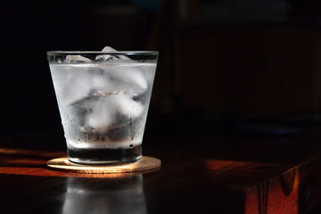 Glass of water with ice cubes on wooden table; shades of sunlight through from window.