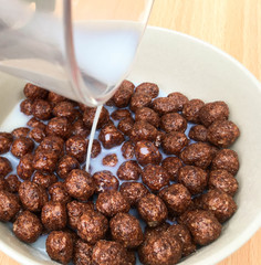 pouring milk into balls of chocolate cereal in a white breakfast bowl on a wooden table,