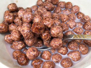 bowl with chocolate balls and a full spoon of chocolate cereal on a wooden breakfast table. Sweet cocoa, chocolate, sugar, cereal, puffs with milk.