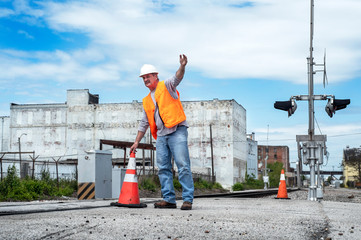 Worker in hard hat and orange safety vest places traffic cone next to railroad tracks train...