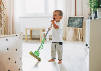 Cute baby boy with mop cleaning floor at room. Child wipes water in the bright interior