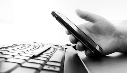 Man's hand holds a smartphone over a black computer keyboard. Heavily blurred background. Black and...
