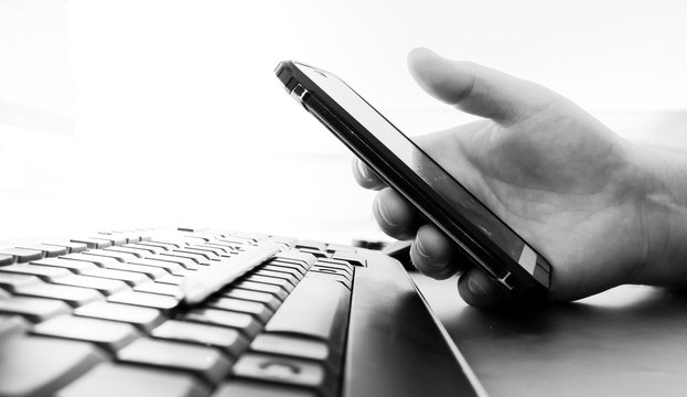 Man's hand holds a smartphone over a black computer keyboard. Heavily blurred background. Black and white image. Copy space.