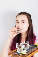 Young girl with pink hair drinks shot of tequila and holds in hand wooden tray with limes, salt and shots on grey background