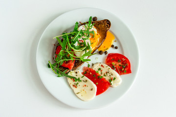 delicious poached egg on a slice of black rye bread with slices of tomato, mozzarella, arugula, spices and black pepper on a white plate on a white background