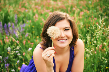 Summer portrait of happy young woman relaxing in wild flower field, playing with dandelion