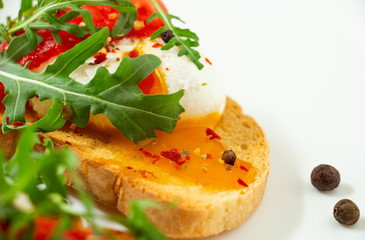 delicious poached eggs on crispy slices of wheat bread with slices of tomato, arugula, spices and black pepper on a white plate on a white background
