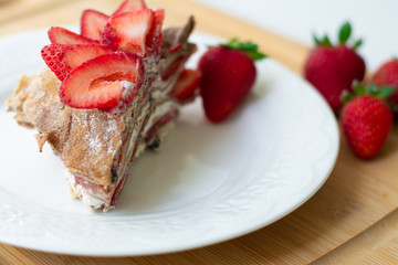 Light summer dessert: a slice of crepe layer cake with whipped cream, chocolate chips and strawberries between the layers. It is decorated with strawberries and placed on a white plate.