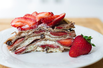 Light summer dessert: a slice of crepe layer cake with whipped cream, chocolate chips and strawberries between the layers. It is decorated with strawberries and placed on a white plate.