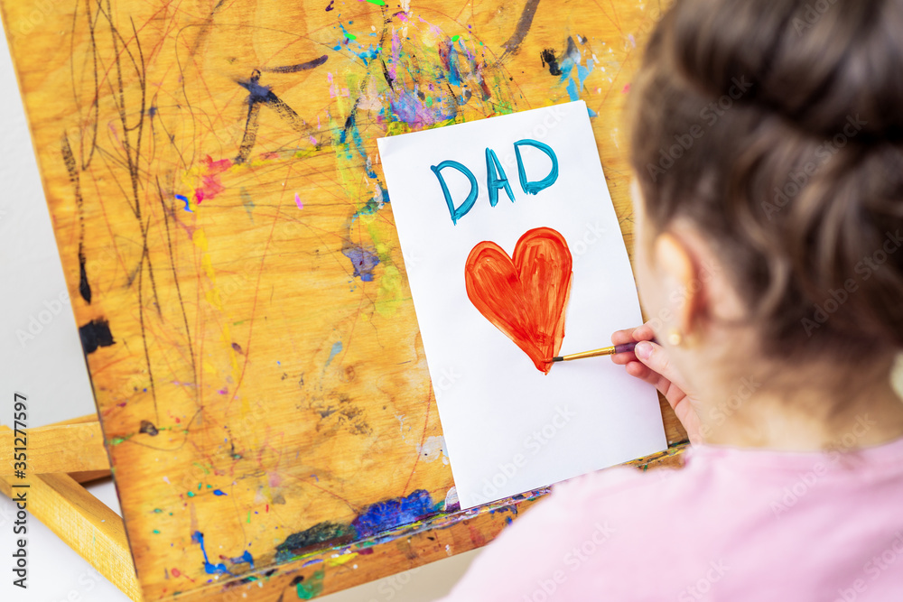 Wall mural Child's hand drawing red heart with word Dad greeting card on white paper on an easel. Happy Father's Day concept.