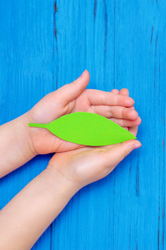 Top view of green paper leaf in hands of child on wooden blue background. Copy space. Concept of environmental protection and Earth day.