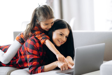 Fototapeta premium Online shopping. Joyful and pretty caucasian mom and daughter are shopping online at home on the couch using a laptop. Nice time together at home