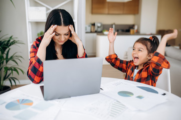 Work at home. Tired young mom try to work remotely on laptop at home, and her cute funny daughter...