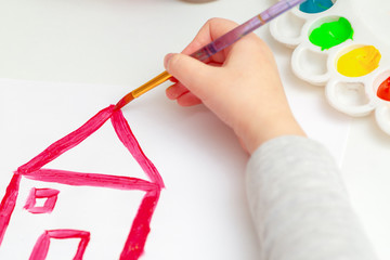 Close up of hand ofn child drawing red house on white paper by watercolors.