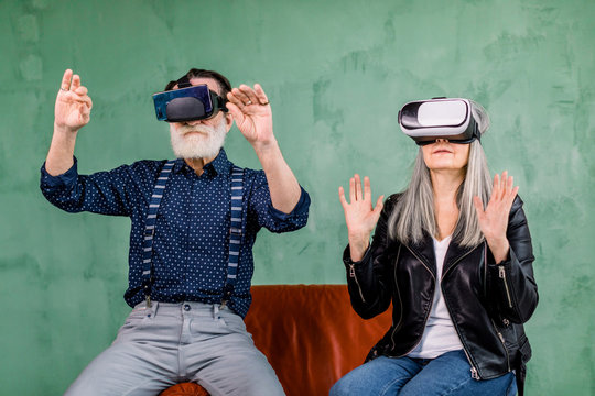 Close up portrait of stylish modern elderly man and woman, wearing trendy clothes, sitting on red chair in virtual reality goggles and touching imaginary screen in the air