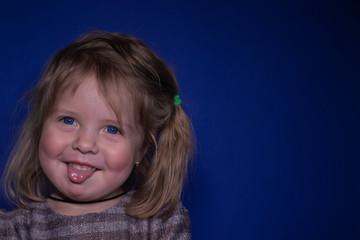 portrait of happy a three year old blond girl with blue eyes showing her tongue on a blue background