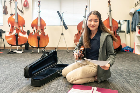 Portrait of smiling teenage girl with violin and sheet music kneeling in classroom