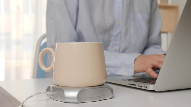 Cup of coffee with heater on table of young woman working with laptop in office