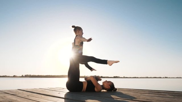 Kids acroyoga performed by two focused acrobats doing support position, pair exercise near water on boardwalk outdoor. Physical activity, practice sport, fitness training together for good body health