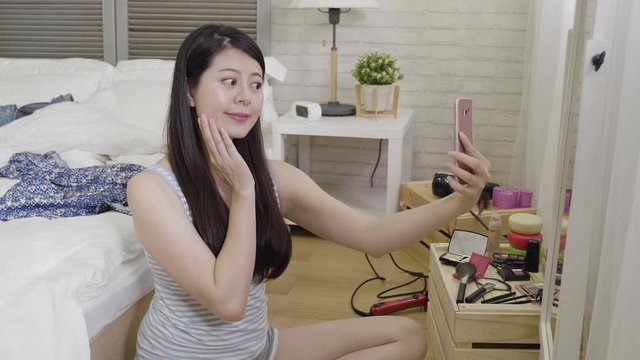 Young beautiful asian woman get ready sitting in on bedroom floor making face smiling taking selfie in the morning. elegant lady using smart phone make self photo in front of mirror enjoy beauty.