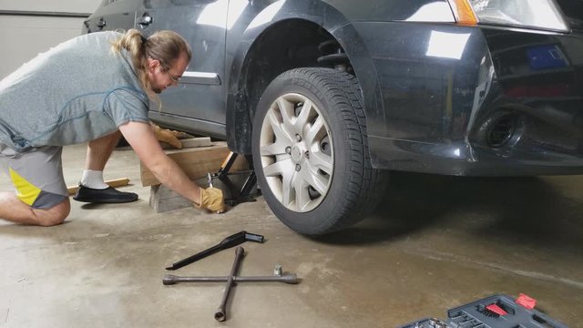 Man in glasses and ponytail wearing shorts use hand jack to rise passenger car while using wood logs supports to stabilize car for later repair or maintenance.