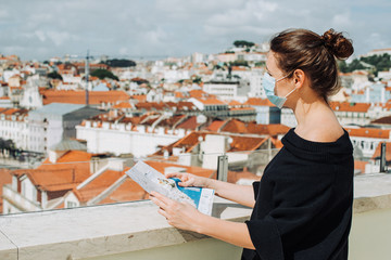 Fototapeta na wymiar Female caucasian tourist wearing a protective mask while standing with a map looking at the city view in Lisbon, Portugal. New normal concept, travel destination or crisis in tourism concept