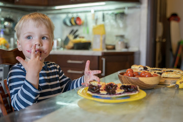 Little cute blond boy licks sweet fingers while eating delicious homemade berry pie in kitchen. Tasty food for kids