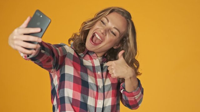 Young cheerful girl isolated over yellow wall background take a selfie by mobile phone shoeing peace