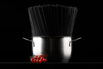 Black spaghetti pasta in a saucepan with fresh tomatoes on a black background.