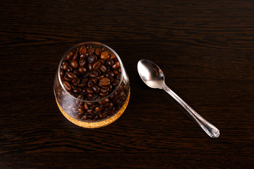 Coffee beans in a glass goblet isolated on a wooden background. Glass on a stand made of balsa wood. A teaspoon lies on the table.