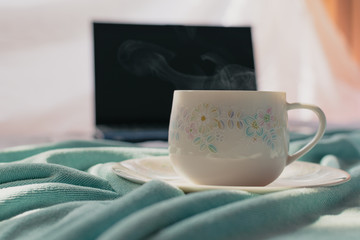 A cup of tea on the bed and light blue blanket with sunrise, Morning teatime with notebook computer and smart phone, Normal practice by working at home during covid-19 outbreak around the world.