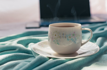 A cup of tea on the bed and light blue blanket with sunrise, Morning teatime with notebook computer and smart phone, Normal practice by working at home during covid-19 outbreak around the world.