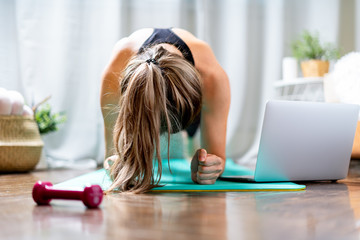 Young fitness woman working out and using laptop and dumbbells at home in living room, doing yoga or pilates exercise on turquoise mat, standing in plank pose. Sport and recreation concept.