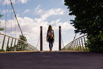 Blonde girl with sunglasses, cap and gold jacket walking across the red bridge at sunrise. River bank. Lifestyle concept and freedom.