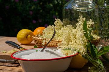 On the wooden table there is an enamel bowl with sugar, a vanilla pod and a basket with fish harvested elderberry blossoms to make a liqueur