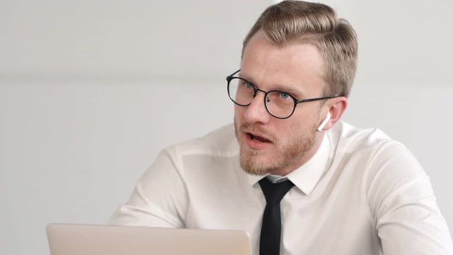 Portrait of young bearded businessman is talking in work process at table in modern office. Successful european entrepreneur speaking and looking at laptop screen while sitting in light workspace