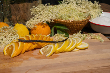 On a cutting board lie a sliced organic lemon and organic orange and a vanilla pod for the elderflower liqueur in the background are elderflowers