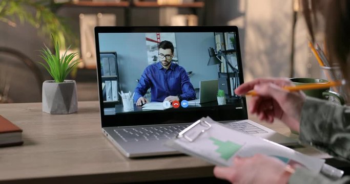 Female worker having business video conference with male boss and writing down information in cabinet. Woman with graphics and pencil taking notes while talking on video call with Caucasian man