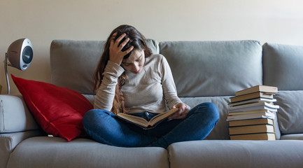 reading a novel on the sofa at home, next to a red cushion and a pile of books