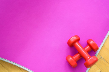Top view of fitness equipment - pink mat for workout, two red dumbbells, on wooden floor. Copy space.