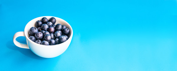 Ripe blueberry and green leaves in cup on blue horizontal background with copy space for breakfast