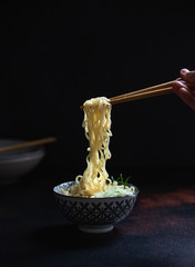 Asian ramen noodle soup with mushrooms, cabbage, green onions and egg in ceramic bowl. Hand holding chopstick with noodles. Japanese street food. Dark background, close up, copy space